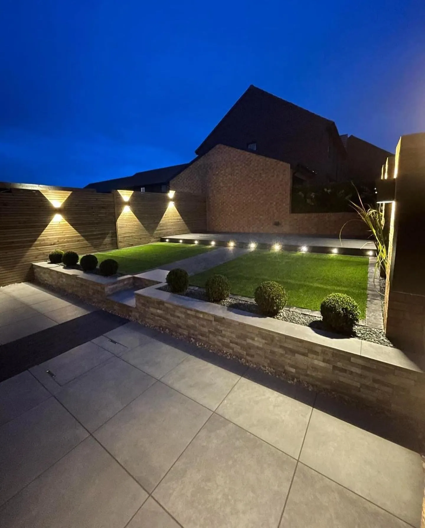 A porcelain patio and new lawn lit at dusk, built by De Builder and Landscape Ltd